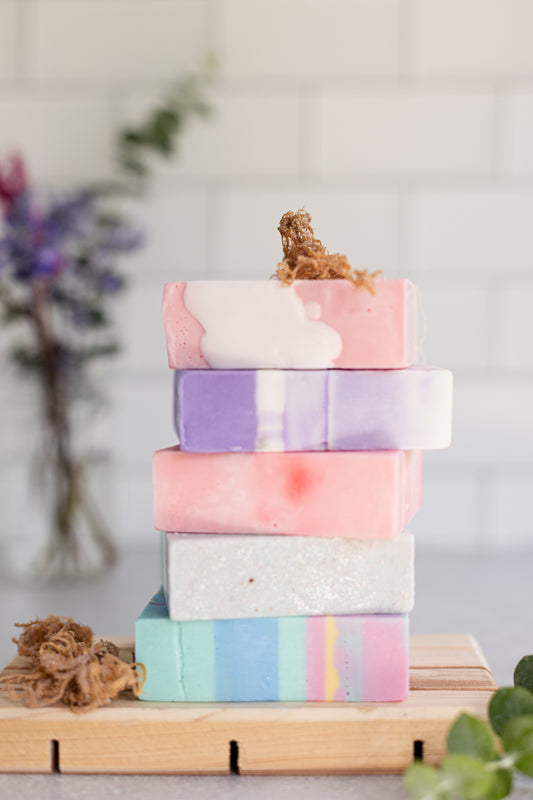 Stack of handmade Inherent Sweetness sea moss soaps on a wooden tray, with the pink and white 1976 Pink Lady bar on top, symbolizing breast-cancer awareness and the friendship that inspired the brand.